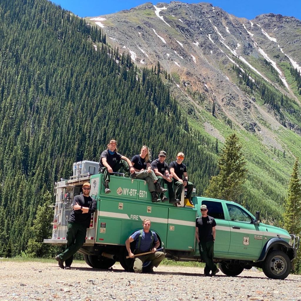 "Photo of Engine 671 with some crew members sitting on top and some standing in front. Steep rocky mountain in background, hill slope covered in pine trees"