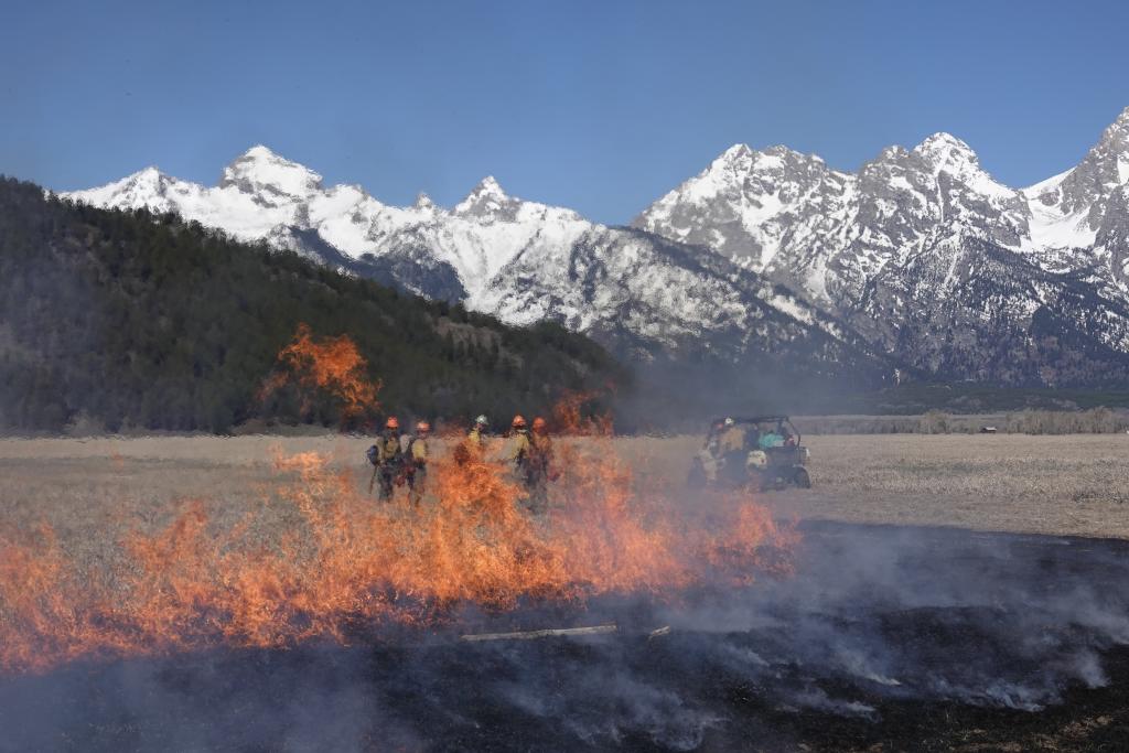 Line of flames with smoke on one side and dry grass on the other with firefighters and Grand Teton mountain in the background