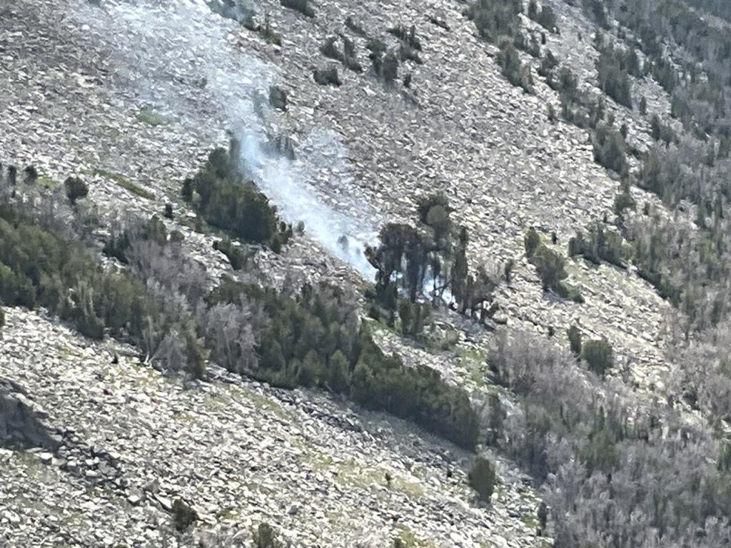 image of the Mount Nystrom fire burning in small clump of trees surrounded by rocks, small amount of white smoke drifting from fire